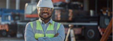 Smiling contractor wearing a safety hardhat, goggles, and vest stands outside a commercial construction site.