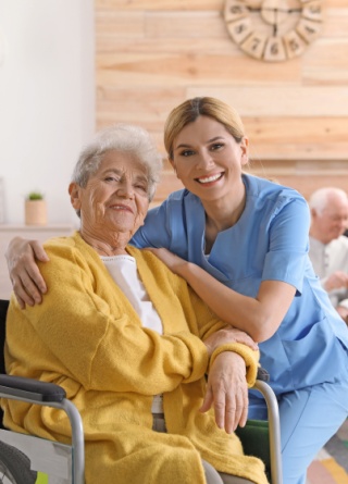 Nurse and woman in wheelchair smile, while other residents and nurse in assisted living facility living room are behind them.Â 