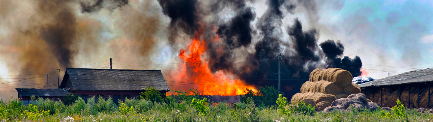 A farm on fire with black smoke next to haybales 