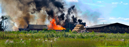 A farm on fire with black smoke next to haybales 