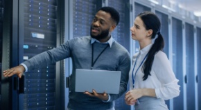 IT Technician with a Laptop Computer points out the server racks to a coworker. 