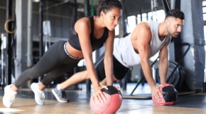 Young man and woman exercising with medicine ball in gym, looking ahead; gym equipment is blurred in the background.