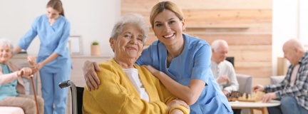 Nurse and woman in wheelchair smile, while other residents and nurse in assisted living facility living room are behind them. 