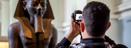 Tourist using conventional camera takes photo of Egyptian statue in a museum as he wonders about museum exhibition insurance. 