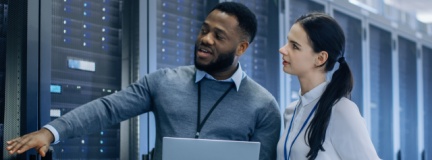 IT Technician with a Laptop Computer points out the server racks to a coworker. 