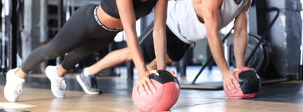 Young man and woman exercising with medicine ball in gym, looking ahead; gym equipment is blurred in the background.